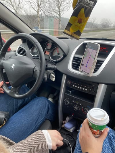 The image depicts the interior of a black Peugeot vehicle, viewed from the front passenger seat, while in motion. A driver, identifiable by hands on the steering wheel and legs in blue jeans operating the pedals, is present. The speedometer indicates approximately 60-70 km/h. A central dashboard display shows "11:14" and "19.9 C." A smartphone, housed in a clear, crystal-embellished mount, is attached to the passenger-side air vent, displaying a navigation map with "57 km" remaining and the time "11:14." A black air freshener with a yellow orchid and "AREON VANILLA BLACK" text hangs from the rearview mirror area. Keys, including a white fob, are in the ignition. The passenger's right hand holds a white disposable coffee cup with a green and red holiday-themed design, resting on a blue knitted item. The passenger's left hand holds a white charging cable, which leads to a black bag between the seats, near a purple earbud case and a white tissue pack. Outside the vehicle, a paved road, bare deciduous trees, and distant hills are visible under an overcast sky. A blurred object displaying green, white, and red stripes (colors of the Bulgarian flag) is visible beyond the driver's side of the dashboard. The scene is located in or near Ushintsi, Bulgaria.