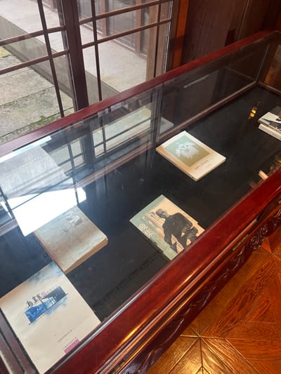 An interior scene in Tongxiang, China, captures a dark wooden display case with a glass top, set against a multi-pane wooden window overlooking an outdoor area. The display case's front border features carved ornamentation. Inside, four books are horizontally arranged on a dark surface. From right to left and top to bottom, these include a light-colored book with a green and white graphic, a light-colored book featuring a black and white photograph of a man in a dark jacket with vertical text, an older, aged brown book, and a white book with a blue and white graphic of a building over water. The glass top reflects the interior, causing some visual obstruction. Outside the window, a paved courtyard with visible moss is present, along with another structure featuring similar wooden window frames. The interior floor in the bottom right is patterned with polished wooden planks.