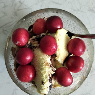 A close-up, top-down shot shows a clear glass bowl filled with ice cream and topped with bright red cherries. The ice cream appears to be a swirl of vanilla and chocolate flavors, with streaks of chocolate sauce drizzled throughout. There are at least eight cherries visible, their smooth, round surfaces reflecting the light. The bowl sits on a light-colored, speckled surface, possibly a marble countertop. A silver spoon is partially visible in the upper right corner of the bowl, suggesting the ice cream is ready to be eaten. The overall impression is one of a simple, refreshing dessert. There is no visible text in the image.