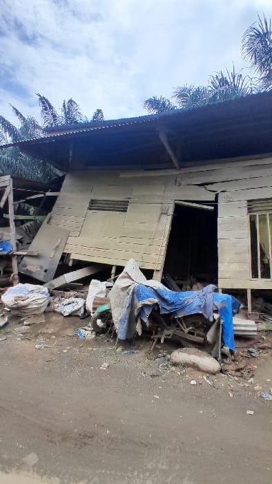An old, weathered wooden house is situated on a dirt road. The house appears dilapidated, with some damage or disrepair visible. There's debris scattered near the house, including what seems like blue and white tarp. Palm trees are visible in the upper left corner of the image, against a cloudy sky. The image has been rotated.