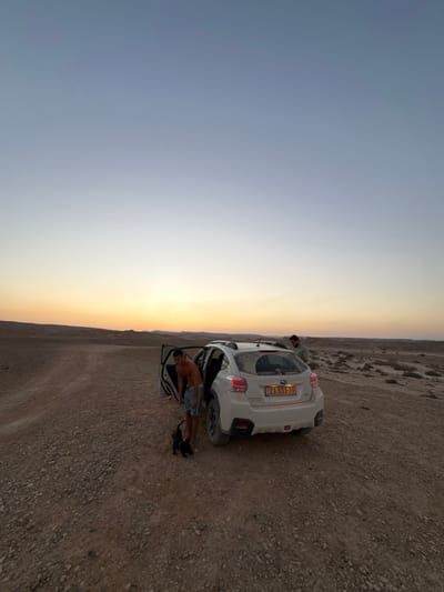The image depicts a desert environment within the Arava Tichona Regional Council, Israel, at either dusk or dawn. A white Subaru Crosstrek, visibly dusty, is stationary on a reddish-brown gravel road. The vehicle's driver's side door is open, and its Israeli license plate, "23-533-39", is legible on the rear.
Two adult males are present. One male, shirtless and wearing light-colored shorts, is partially exiting the open driver's side of the vehicle, appearing to be bending or reaching inside. A small, dark-furred dog stands on the ground beside him, near a dark bag. A second male, wearing a long-sleeved shirt, stands on the right side of the vehicle, near its rear passenger area.
The surrounding terrain is characterized by barren, rocky ground with sparse, low-lying scrub vegetation in the middle distance. Low hills or elevated desert formations are visible on the horizon. The sky exhibits a gradient from a clear, deep blue at the top, transitioning to light blue, and then to a distinct yellow-orange hue at the horizon.