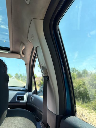 The image shows the interior of a car, specifically the roof and part of the dashboard. The car has a sunroof. Outside the car, vegetation can be seen. The sky is blue and clear. The location is likely outdoors, as evidenced by the natural light and vegetation. There are no discernible people or actions taking place, beyond the implicit fact of someone being in the car. The location of Belovitsa, Bulgaria is not evident from the image.