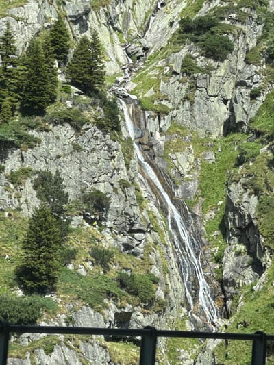 The image depicts a steep, rocky mountainside with a prominent waterfall cascading down its face. The terrain is characterized by large, grey rock formations interspersed with patches of green vegetation, including trees and grass, indicating a natural, mountainous environment. The waterfall itself is a series of white streams of water flowing over dark, wet rock. The scene appears to be an outdoor, wilderness setting, likely in a mountainous region. Given the location of Andermatt, Switzerland, this is likely a view of one of the many natural attractions near the town, possibly a gorge or a mountain stream. No people, animals, or man-made structures other than a dark railing in the foreground are visible.