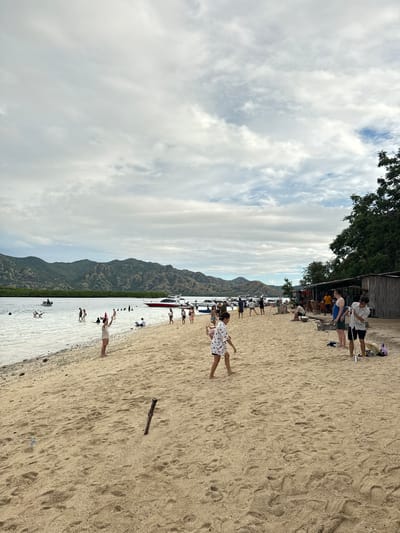 The image depicts a beach scene in Rinca Village, Indonesia. The foreground features a sandy beach with multiple footprints and a piece of driftwood. People are scattered across the scene, some wading in the water and others standing on the sand. In the midground, several boats are docked near the shore. In the background, there are green mountains under a cloudy sky. At the top left corner of the image, there are trees and a building.