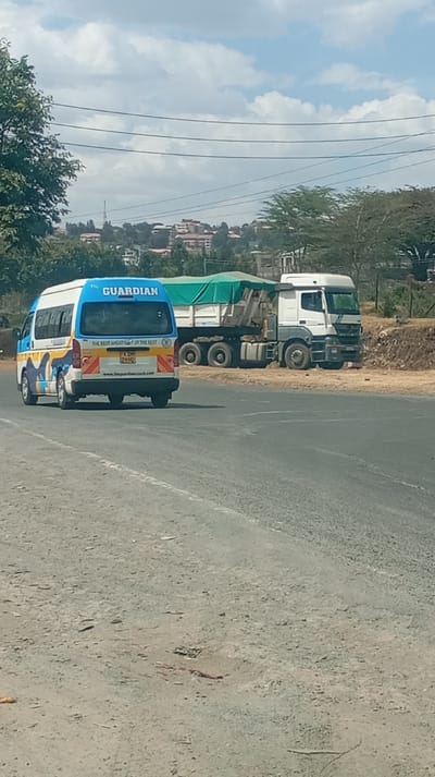 A road stretches towards a background of buildings and sky. A blue and white van labeled "The Guardian" is on the left. To the right, a large white truck with a green-covered trailer sits stationary beside the road. Trees dot the horizon behind the truck. Utility wires are strung above the scene.