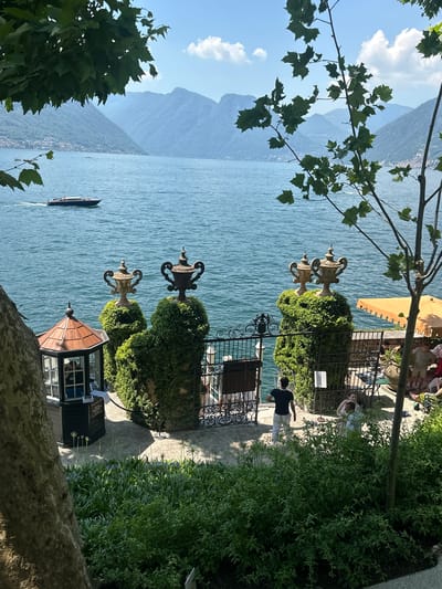 The image depicts a scenic view of Lake Como in Tremezzina, Italy. In the foreground, lush green foliage frames the scene, with a large tree trunk on the left and branches overhead. A small, ornate gazebo with a copper roof is visible on the left, beside a black structure with windows, possibly a ticket booth or information kiosk.
Dominating the middle ground are two elaborately trimmed topiaries shaped like urns, each topped with a decorative urn statue. Between them is an open wrought-iron gate, with more ornate fencing to the right. Beyond the gate, a paved area is visible, with a few people seated at tables under a yellow umbrella.
The water of Lake Como stretches into the background, with a boat cruising across its surface. In the distance, rolling mountains rise, covered in verdant vegetation and dotted with buildings, suggesting the presence of villages along the shore. The sky is a clear blue with a few wispy clouds, indicative of a sunny day.
Several people are present in the image. A man in a black t-shirt and white pants stands near the gate, facing away from the camera. A woman and a child are visible in the background, near the seated patrons. Another person is partially visible inside the gazebo. The overall scene conveys a serene and picturesque atmosphere of a lakeside garden estate.
