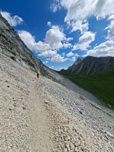 An extreme wide shot captures a lone individual traversing a rugged, rocky path on a mountain slope. The path is a narrow, light brown strip winding through a vast expanse of grey and tan scree. To the left, a steep, grey mountain face rises dramatically, its surface textured with rock and sparse green vegetation. To the right, the slope transitions into a vibrant green meadow that stretches towards a distant, hazy mountain range. The sky above is a brilliant blue, dotted with fluffy white clouds. The person, small in the frame, is walking away from the camera, their dark clothing providing a stark contrast to the muted tones of the terrain. The overall scene depicts a solitary hike in a vast, natural landscape, suggesting an environment found in mountainous regions like the Austrian Alps near Mieming.