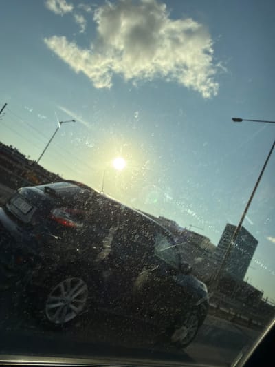 The image appears to be taken from inside a vehicle, looking out through a dirty windshield. The sun is bright and appears to be low in the sky, causing glare. In the foreground, the back of a dark-colored car is visible, with its taillights illuminated. The car has a license plate with some visible characters. In the background, there are several buildings, possibly skyscrapers, and streetlights. There are also some power lines or other infrastructure. The sky is blue with a few clouds. The overall impression is of a daytime scene in a city. The image is hazy and out of focus, which could be due to the dirt on the windshield, camera settings, or motion blur. There are no people clearly visible in the image.
The prompt mentions Mölndal, Sweden, but there are no visual cues in the image that specifically identify this location.