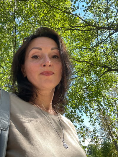 A medium shot, eye-level, outdoor selfie shows a middle-aged woman in the foreground, looking directly at the camera. She has dark, shoulder-length hair, brown eyes, and is wearing light-colored clothing with a silver necklace. She appears to be in a park or natural setting, with lush green trees and a blue sky visible behind her. The lighting suggests it is daytime. The specific location in Kirovo-Chepetsk, Russia, is not discernible from the image.