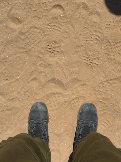 The image shows a pair of dark gray combat boots standing on light brown, sandy ground. The boots are laced with black laces and appear worn. The sandy ground has numerous footprints and impressions, indicating recent activity. The lighting suggests a sunny outdoor environment. The bottom of the image shows the lower legs of a person wearing olive green pants, partially obscuring the boots. The scene is consistent with an outdoor, arid or semi-arid location. The location is Bnei Shimon Regional Council, Israel, which is known for its desert and semi-arid landscapes.