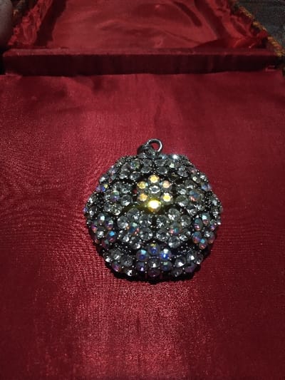 A circular, domed piece of jewelry, possibly a pendant or brooch, is centrally positioned on a dark red, satin-like fabric. The object is heavily embellished with numerous small, clear, faceted stones, likely rhinestones or crystals, set into a dark, possibly silver-toned or black metal base. These stones exhibit iridescent reflections, primarily blue, pink, and white, with a notably brighter, yellowish glow emanating from a cluster of stones near the center-top of the object. The fabric surface shows minor creases and a subtle sheen. In the upper portion of the image, a raised, upholstered edge consistent with the interior lining of a presentation box or display case is visible, also covered in the same dark red fabric. The lighting is dim, emphasizing reflections from the stones. No people are visible. No specific location within the city of Domažlice, Czechia, is identifiable from the visual evidence provided.