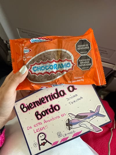 A person is holding a Chocoramo snack and a welcome card. The snack is in an orange wrapper with the words "CHOCORAMO" in a red and white logo. The wrapper also has warning labels regarding excess sugar and saturated fats, in Spanish. The welcome card is light pink and decorated with dots. It has the words "Bienvenida a Bordo" (Welcome Aboard) in large letters, along with the names "Jessica" and "Takuma", and the flight number "LA4406". The card also has a sticker of a cartoon airplane. The person's hand with a light blue nail polish is visible. The background appears to be the interior of an airplane. The image does not depict any location in the city of Orlando, Florida, United States of America.