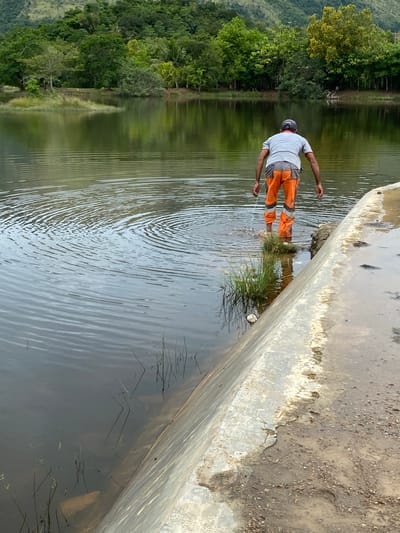 In Aneuk Paya, Indonesia, a person wearing a grey shirt, a dark baseball cap, and orange work pants with reflective stripes stands in shallow water near a concrete structure. The individual is facing away from the camera, with their hands lowered towards the water's surface, creating ripples. The environment consists of a body of water reflecting a lush green hillside covered in trees. The concrete structure appears to be a dam or retaining wall, with a rough, textured surface and some debris on the adjacent dry ground. The water is calm, with visible concentric ripples originating from the person's movement.