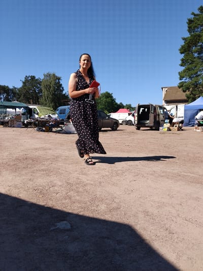 A woman in a black polka-dot dress is walking in an outdoor market on a sunny day. She is holding a red clutch and a glass. In the background, there are tents, vans, and trees. The ground is covered in dirt and gravel. The location is identified as Irshansk, Ukraine.