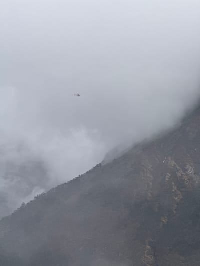 The image captures a dramatic scene of a helicopter navigating through dense fog or clouds in a mountainous region, specifically the Namche Bazaar area of Nepal. The majority of the frame is filled with a thick, ethereal expanse of greyish-white fog, creating an atmosphere of low visibility and vastness.
A small, distinct helicopter is visible in the upper-left quadrant of the image, appearing somewhat isolated amidst the heavy mist. It has a light-colored body with subtle darker accents or markings on its tail and rotor mechanism. The helicopter's presence suggests activity at high altitude, likely flying through challenging weather conditions.
To the right side of the frame, a dark and rugged mountain or cliff face partially emerges from the swirling clouds. Its surface is characterized by rough, stony textures in shades of dark grey and brown, indicating a barren, rocky landscape. The abrupt edge where the mountain meets the fog creates a stark visual boundary.
The weather is clearly very foggy or cloudy, obscuring distant views and enveloping the terrain. The time of day is difficult to ascertain precisely due to the pervasive cloud cover, but the diffused lighting suggests daytime. No discernible text is visible within the image. The overall impression is one of grandeur, isolation, and the formidable nature of the Himalayan environment.