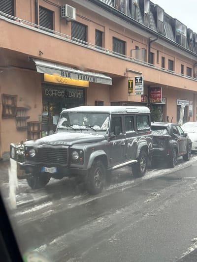 The image depicts a street scene in Lipomo, Italy, during daylight hours under snowy conditions. A dark grey four-door Land Rover Defender (license plate ZA 781TE) is parked on the right side of a street, facing left. The vehicle is coated with a light layer of snow, exhibiting decorative patterns on its side doors and roof. Behind the Land Rover, a dark-colored SUV, also partially covered in snow, is parked. The road and visible pavement areas are covered in slushy snow and ice, with tire tracks present.
To the left of the vehicles, a two-story building with a light orange-peach facade stands. The ground floor houses commercial establishments. A "BAR TABACCHI" with a beige awning and a yellow illuminated sign displaying "BAR" and "COFFEE & DRINKS" is prominent. To its right, another sign with a large "T" indicates a "TABACCHI" and "BAR." Further right, a sign for "IL FARAONE PIZZA & KEBAB D'ASPORTO" is visible. The second story of the building features windows with dark roller shutters and an external air conditioning unit above the "BAR TABACCHI." The roofline above the second story includes multiple dormer windows. No individuals are visible.