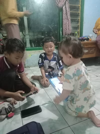 The photograph captures a scene indoors, likely in a home located in Bungur, Indonesia. Three children are the central figures of the image. Two boys are seated on the floor, both engaged with electronic devices, presumably smartphones or tablets. The boy on the left is wearing a maroon collared shirt and dark pants, and he is focused on a phone in his hands. Beside him is a plush toy. The boy seated next to him is wearing a jersey with a number 3 and a graphic print. He is holding a tablet. In front of him, is a young child, possibly a toddler, wearing a light-colored outfit with a pattern of dinosaurs. They are also holding a tablet. The floor is tiled with light-colored square tiles. In the background, an adult is visible. The photograph suggests a casual, domestic setting.