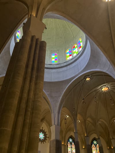 The image presents an upward-looking view inside a monumental architectural space, showcasing its intricate ceiling and upper structural elements. The scene is dominated by massive, smooth, and organically shaped columns and vaulted ceilings, likely constructed from concrete or light-colored stone. These structural elements branch out and converge, creating a complex, tree-like canopy effect overhead.
Several stained glass windows illuminate the interior. On the left side of the frame, a series of rectangular windows feature a modern, abstract design with bright, distinct blocks of primary and secondary colors (red, green, blue, yellow, magenta, cyan, white) arranged in a grid. Further into the distance and towards the right, other circular or rosette-shaped stained glass windows display more traditional, ornate patterns with a vibrant mix of colors.
Small, warm-toned light fixtures are subtly integrated into the ceiling vaults and along the structural arches, contributing to the overall subdued and majestic illumination. The architecture is characterized by sweeping curves, geometric precision, and a blend of modern and organic forms, creating a sense of grandeur and awe.
There are no people or specific activities depicted; the focus is entirely on the building's interior. The overall lighting is warm, suggesting an ambient or artificial light source complementing any natural light filtering through the stained glass. No visible text is present in the image. The location context provided is San Cristóbal De La Laguna, Spain.
