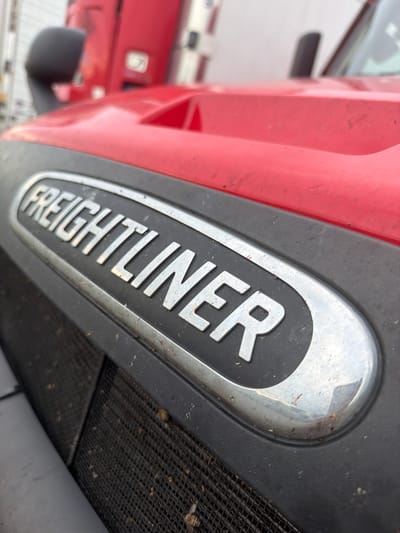 The image is a close-up, low-angle, outdoor photograph of the front grille and hood emblem of a red Freightliner semi-truck. The emblem, which reads "FREIGHTLINER" in silver raised letters within a chrome-finished oval, is the central focus. The truck appears to be parked, possibly in a lot or on a street in Murfreesboro, United States of America. The foreground shows a black mesh grille, partially obscured by the hood. The red paint of the hood is visible, showing some dust and minor scuffs. The background is slightly blurred, showing more of the truck's cab and possibly other vehicles or structures. The lighting suggests it is daytime. There are no people visible in the image.