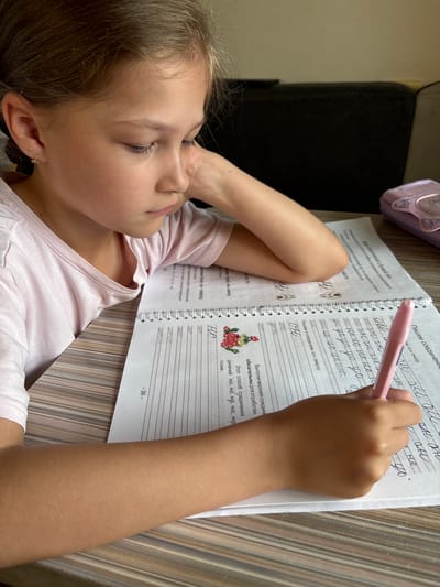 The image depicts a young girl, appearing to be of Eastern European descent, engaged in writing in a notebook. She is positioned at a table, with her left arm resting on it and her head tilted downwards as she concentrates on her work. She is wearing a light pink t-shirt. In the foreground, a pink pen is held by her right hand, poised over the notebook. The notebook contains handwritten text and line guides, suggesting it is an educational workbook, possibly for practicing calligraphy or penmanship. The environment appears to be a domestic setting, with a dark, upholstered object visible in the background, possibly a sofa or chair. A light purple pencil case, adorned with a cat's face design, is also present on the table. The lighting suggests an indoor setting, potentially during daylight hours. The girl's facial expression indicates focus and perhaps a slight furrowing of her brow. The table surface has a wood-grain pattern. There is no visible indication of the specific location being Bratsk, Russia.