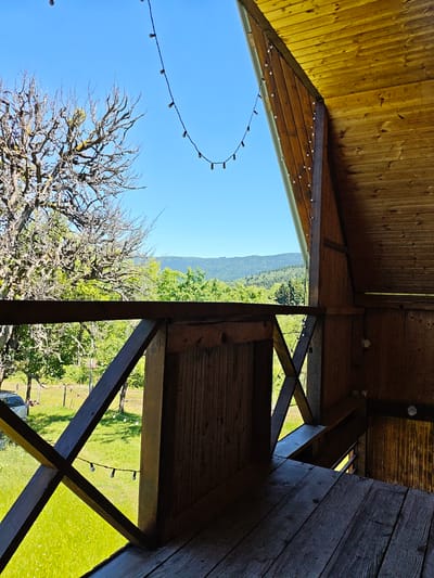 The image captures a view from a wooden balcony overlooking a lush green landscape under a clear blue sky. In the foreground, weathered wooden planks form the balcony floor, and a dark wooden railing with an X-shaped support structure divides the view. String lights, composed of small white bulbs, are draped across the scene, some hanging from the balcony roof and others across the railing.
To the left, a large, bare tree with gnarled branches stands against the sky, its silhouette contrasting with the vibrant green foliage of smaller trees and shrubs in the middle ground. Beyond the trees, rolling hills covered in dense forest stretch towards the horizon. The overall impression is one of a serene, natural setting. The scene is devoid of any people or animals. The location is identified as Ukeshi, Georgia, suggesting this balcony offers a vantage point for observing the region's natural beauty.