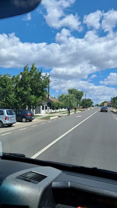 The image depicts a street scene in Balș, Romania, on a clear day with scattered clouds. The perspective is from inside a vehicle, looking out through the windshield and dashboard. On the left side of the road, there are two parked cars: a silver minivan and a dark blue hatchback. Adjacent to the parked cars is a sidewalk and a building with a white facade and a dark roof, surrounded by lush green trees. Further down the road, two individuals are cycling. The cyclist on the left is wearing a blue shirt and black shorts, while the cyclist on the right is wearing a black t-shirt and shorts. They are riding in the same direction as the camera vehicle. On the right side of the road, there are more trees and houses in the background, with power lines visible overhead. A dark-colored car is traveling in the distance on the right lane of the road. A speed limit sign with "50" inside a red circle is visible on the road in front of the cyclists. The road is paved and has white lane markings.
