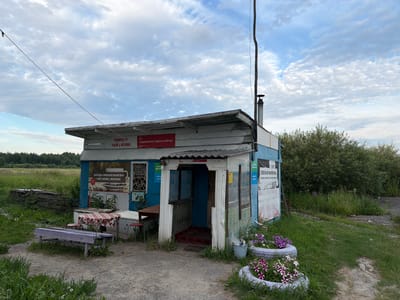 The image depicts a small, rustic roadside shop or kiosk in the city of Alga, Russia. The building is constructed of weathered wood and metal, with a covered porch area leading to the entrance. A sign above the entrance reads "ПИРОГИ ЧАЙ | КОФЕ" (Pies Tea | Coffee) and another sign advertises "ПОДКЛЮЧИ МТС ЗДЕСЬ И СЕЙЧАС!" (Connect MTS Here and Now!).
Outside the building, there is a red and white checkered tablecloth on a table, with two red stools. A purple wooden bench sits to the left of the table. In front of the building, there are two car tires repurposed as planters, filled with purple and white flowers. The surrounding environment is rural, with tall green grass, a dirt path, and dense green trees in the background. The sky is partly cloudy with a mix of blue and white. There are no people visible in the image. The scene suggests a simple, functional establishment catering to travelers or locals in a somewhat remote area.