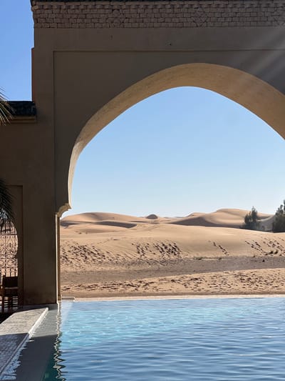 An outdoor scene in Rissani, Morocco, is viewed through a light-brown stucco archway. The lower foreground features a clear blue swimming pool with visible ripples and light-colored tiled edges, including steps on the left. The archway, constructed of a beige material and adorned with decorative brickwork along its upper edge, frames a distant desert landscape. To the immediate left of the arch, a section of a building wall, a dark, ornate metal gate with intricate patterns, and a single palm frond are partially visible. Beyond the arch, a vast expanse of golden-brown sand dunes stretches under a cloudless, bright blue sky. The sandy terrain exhibits numerous human footprints and vehicle tracks. Several sparse green trees are present on the far right within the dune field. The environment is brightly illuminated by natural sunlight, casting shadows under the arch. No people are visible.