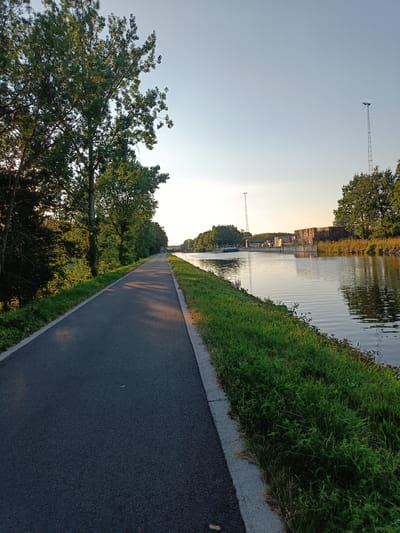 The image depicts a paved path running alongside a tranquil canal. Trees line the left side of the path, casting dappled shadows on the asphalt. The canal, on the right, reflects the clear sky and the lush greenery of its banks. In the distance, a barge is visible on the water, and industrial structures, including stacks of pallets and a tall lighting tower, are situated on the far bank. The scene appears to be set in Haacht, Belgium, likely along the Leuven-Dijle Canal, a waterway that flows through the region. The overall atmosphere is peaceful, suggesting a serene natural environment that is also utilized for industrial transport.