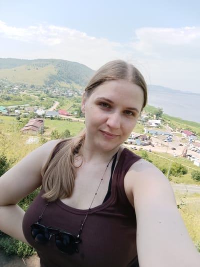A young Caucasian woman with light brown hair pulled back into a ponytail takes a selfie outdoors in the city of Solnechnaya Polyana, Russia. She is wearing a dark brown tank top with sunglasses hanging from a chain around her neck. In the background, a village with scattered houses and buildings is visible, nestled in a valley. A large body of water, likely a river or lake, extends to the right of the frame, with a distant hilly landscape on the horizon. The weather appears to be clear and sunny, with a few scattered clouds in the sky.