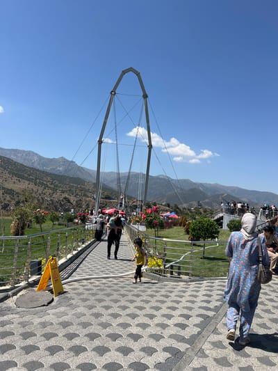 The image depicts a sunny outdoor scene at the Dardar Gorge in Uzbekistan, featuring a modern pedestrian bridge. The bridge's structure is characterized by a large, arched metal frame supported by cables, which stretches across a mountainous landscape. A patterned walkway, paved with grey and black interlocking semi-circular tiles, leads across the bridge.
Several people are visible on and around the bridge. In the foreground, a child in a yellow top and black pants walks along the path. A woman wearing a blue patterned tunic and pants, with a headscarf, walks away from the camera on the right side of the walkway. Further down the bridge, a man in black clothing carries a child. Other individuals and groups are scattered across the bridge, some looking at the scenery or interacting with each other.
The surrounding environment includes lush green grass on either side of the walkway, with decorative flowers and small trees. Beyond the bridge, a mountainous terrain with dry, rocky slopes and sparse vegetation is visible under a clear blue sky with scattered white clouds. Red and blue tents, likely for vendors or recreational activities, are set up in the middle ground. In the distance, more mountains stretch across the horizon. A yellow "Caution Wet Floor" sign is placed on the left side of the walkway near a manhole cover. The overall impression is of a popular tourist spot with a mix of natural beauty and man-made structures.