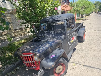 A black, vintage Willys Jeep pickup truck sits parked on a paved street. The vehicle features a red grille, round headlights, and tires with red rims. An exhaust pipe extends upwards from the hood. The driver's side door displays a checkered flag design and writing in white. Trees line the street, and a house with a red-tiled roof is visible in the background.