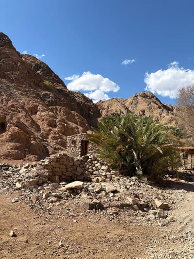 The scene depicts a mountain landscape in Dahab, Egypt. A red-brown rocky mountain occupies the left side of the frame, its textured surface marked by crevices and shadows. A stone structure, possibly a small building or part of a larger complex, is built into the mountainside, featuring a dark-colored window or opening. Adjacent to the structure, a date palm tree with dense green fronds grows from the rocky terrain. The right side of the frame shows a dirt path or road, covered with pebbles and stones, leading towards the mountainside. Above the mountains, a bright blue sky with scattered white clouds is visible.