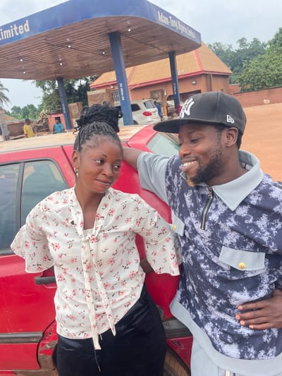 Two people are interacting next to a red car at what appears to be an Adam-Tony Nigeria Limited gas station in Otukpa, Nigeria. A man in a black baseball cap and a patterned shirt is leaning on the car's hood, smiling at a woman in a white floral blouse, who is standing next to the car, looking up at him. They are close together. Behind them, there are buildings, other cars, and people. The environment includes elements of a gas station, such as the sign and pump.