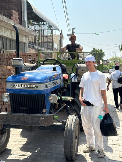 A man with a bandana poses next to a blue Sonalika DI-35 tractor on a brick-paved street in Mathura, India. The tractor is parked in front of stacked bricks. Another man is sitting on the tractor behind him. A man is walking on the side of the street with a black bag and a water bottle.