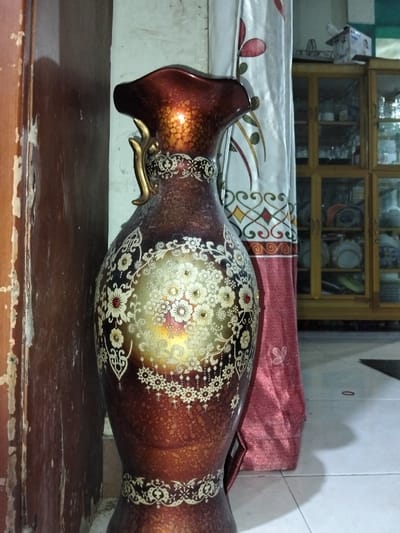 A tall, ornate vase stands prominently. The vase is a blend of maroon and brown hues, adorned with intricate floral and geometric designs in gold and white. The upper portion features a flared lip and a decorative handle. The vase sits on a white tiled floor near a distressed wooden door and a curtain patterned with red and white designs. A wooden cabinet filled with various items is visible in the background.