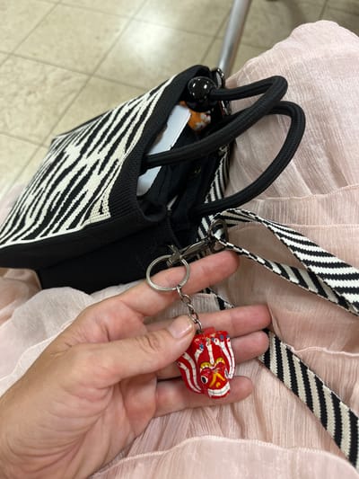 A close-up shot shows a hand holding a small, red, wooden mask-shaped keychain with white, blue, and yellow accents. The keychain is attached to a silver keyring and a carabiner clip, which is clipped onto the black and white striped strap of a black tote bag. The bag has a black and white zebra-print fabric panel on the front and black rope handles. The bag is resting on a light pink, crinkled fabric, possibly a dress or skirt. The background is out of focus but appears to be a tiled floor. The scene suggests a casual setting, possibly a public place like a cafe or a bus stop in Katunayake, Sri Lanka, given the cultural design of the keychain.