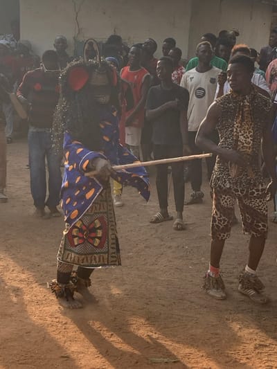 A group of people gathers to watch a cultural performance. In the foreground, a masked figure in a blue and orange robe and adorned with dried vegetation, holds a staff. A person in a leopard-print outfit gestures with their hands, while others observe in casual attire. The scene is bathed in sunlight, casting shadows on the ground.