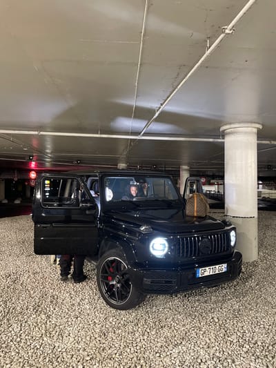 A black Mercedes-Benz G-Wagon with the driver's side door open sits parked in a garage. The car, with a license plate reading "GP-710-GG", is positioned near a grey wall with pipes and a cylindrical pillar. A Louis Vuitton handbag rests on the passenger seat. The driver is visible in the driver's seat. The ground is covered in small, light-colored rocks.