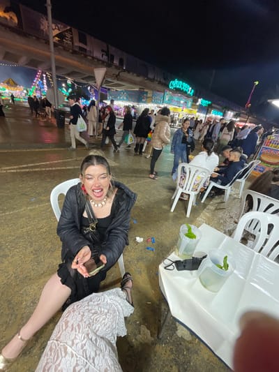 The image captures an outdoor scene at night, likely in Jerez De La Frontera, Spain, given the signage visible in the background. A woman is seated in a white plastic chair, smiling broadly at the camera. She wears a black jacket, a dark top, a pearl necklace, and holds a smartphone. To her right, another person is seated, wearing a white lace dress and gold heels. The ground appears wet, and there are discarded items scattered around. In the background, there is a brightly lit fairground with numerous stalls and a crowd of people. There is a table with water filled cups with leaves next to them and a black face mask.