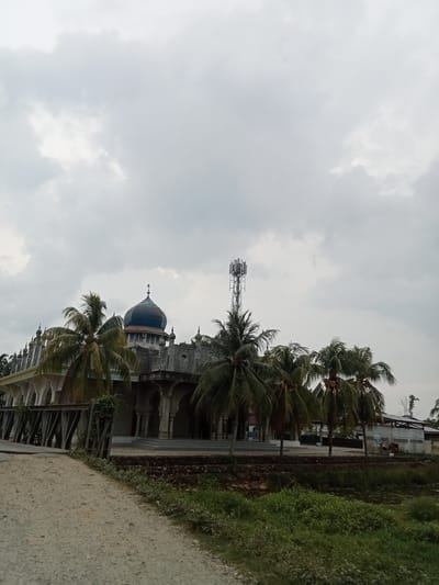 The image depicts a mosque with a blue dome, characterized by intricate architectural details and multiple smaller spires, under a cloudy sky. A cell tower stands tall behind the mosque, surrounded by palm trees. In the foreground, a dirt path leads towards the mosque, with a metallic structure that appears to be a bridge on the left. Lush green vegetation and a body of water are visible to the right of the path. The scene suggests a rural or semi-rural setting in Matangsijuek Barat, Indonesia. No people are visible in the image.