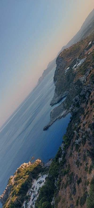 The image displays a coastal landscape with the sea on the left and a rugged, vegetated cliff face on the right. The scene is captured from a high vantage point, looking down and across the coastline.
The water is a deep blue, reflecting the clear sky above, with subtle ripples indicating a calm sea. Several small boats are visible on the water near the shore. The coastline is characterized by rocky outcrops and coves.
The cliff face is covered with dry grasses, shrubs, and scattered trees. On the upper part of the cliff, several white buildings with distinctive architecture, likely resorts or houses, are nestled into the hillside. A winding road or pathway is visible leading up to these structures.
At the base of the cliff on the lower left, a parking lot is visible, filled with numerous cars of various colors. This area is surrounded by dense green vegetation. The overall impression is a sun-drenched Mediterranean coastal scene. The location is identified as Faliraki, Greece.