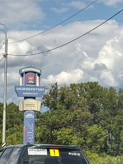 The image depicts a street signpost in Semibratovo, Russia. The signpost is in a vertical orientation and is the main subject. It features multiple signs with text and symbols. The top sign is blue and curved, with text in Cyrillic. Below it is a central sign with a coat of arms and the name "СЕМИБРАТОВО" (Semibratovo) in large blue letters. To the right of this central sign is a golden scroll-like sign, also with Cyrillic text. Another blue sign with Cyrillic text and red and white striped patterns is visible to the right.
The signpost is situated outdoors, with a backdrop of lush green trees and a cloudy sky. Power lines are visible running across the frame. To the right of the signpost, a black vehicle is partially visible, with a "ПРОДАЮ" (For Sale) sticker and a yellow exclamation mark sticker on its rear.
The lighting suggests it is daytime. The weather appears partly cloudy. The overall scene is a roadside view in a rural or semi-rural area. The presence of the "For Sale" sticker on the car hints at a possible transaction or advertisement. The text on the signs indicates the location as Semibratovo, a settlement in Russia.