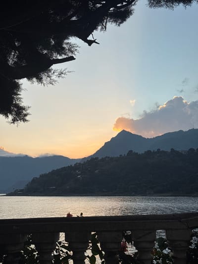 A scenic view features a serene body of water reflecting sunlight. A dark, mountainous landscape dominates the background, partially obscured by a dramatic cloud formation with a bright, sunlit edge. The silhouette of trees frame the left side, contrasting against the lighter sky. A structure, adorned with greenery, occupies the right side of the frame. The photograph is oriented vertically.