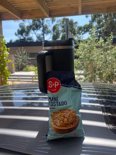 A black insulated mug with a handle, clear lid, and metallic straw stands upright on a reflective, dark gray surface. Resting against the mug is an open light blue bag of "S&P Maní Tostado Sin Sal" (Toasted Peanuts Without Salt), partially filled with peanuts, visible through an image on the bag. The scene is set outdoors in La Barra, Uruguay. A wooden slatted pergola structure is visible overhead, casting distinct parallel shadows across the reflective surface. In the background, under a clear blue sky, are numerous green trees and a modern building with a dark facade. No individuals are present, and the objects appear stationary in bright daylight.