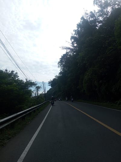 A road, likely a rural highway, stretches into the distance under a cloudy sky. The road has a yellow line marking the center and a white line on the left edge. A guardrail is present on the left side of the road, next to a tree-lined hillside. On the right, a thick forest borders the road. Several people are riding motorcycles along the road, moving away from the camera. The location might be Khe Luang, Laos, although this cannot be confirmed from the visual data.