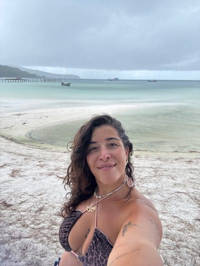 A vertical, selfie-style photo shows a woman on a beach with the ocean in the background. She is wearing a leopard-print bikini and a nameplate necklace. Her hair is dark and wavy, and she has a tattoo on her right shoulder. The beach appears to be sandy with some dried seaweed. The ocean water is a light green color, and the sky is overcast and gray. In the distance, there are a few boats and a pier. The woman is smiling and looking at the camera. The overall mood of the photo is relaxed and happy, suggesting a vacation or a day at the beach. The text visible on the necklace spells out "Maya". The overcast sky suggests it might be a cloudy day or nearing sunset. The location context of Kâmpóng Saôm, Cambodia, implies a tropical or subtropical beach setting.