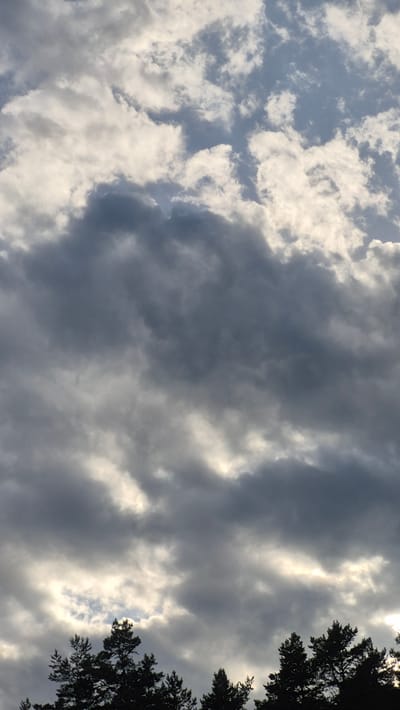 The image shows a cloudy sky above a silhouette of trees. The clouds are varied in texture and density, with some appearing bright white and others dark gray, suggesting a mix of sunlight and shadow. The sky occupies the majority of the frame, with the treetops forming a dark band across the bottom. The scene appears to be outdoors, with no buildings or specific landmarks visible. The location is identified as Ørje, Norway.