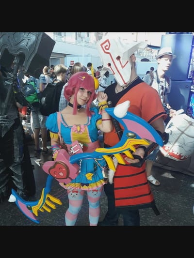 In a crowded indoor space, two cosplayers are the central focus. The individual on the left, dressed as a pink-haired anime character, wears a colorful blue and yellow dress adorned with bows and pink stripes on their legs. They are holding a stylized pink and blue bow with a heart-shaped grip. To the right, another cosplayer sports a white mask with red markings and an orange and red striped tunic. They are holding a large white object with red flames, resembling a drum. The background reveals a diverse crowd of people, some carrying backpacks and wearing casual attire. The environment appears to be an event or convention, with large structures and speakers visible. There is no discernible evidence to suggest the location is the city of Norcia, Italy.