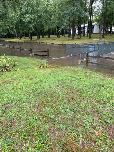 In Hamilton Township, United States of America, a flooded area is visible. A chain-link fence and a wooden fence run through the scene, separating a grassy area from the water. Trees line the background, and a white building with multiple stories is partially visible behind them. The ground is covered in grass, with some areas appearing muddy. Debris, including branches and leaves, is present in the flooded water and along the edge of the grass. The lighting suggests an overcast day.